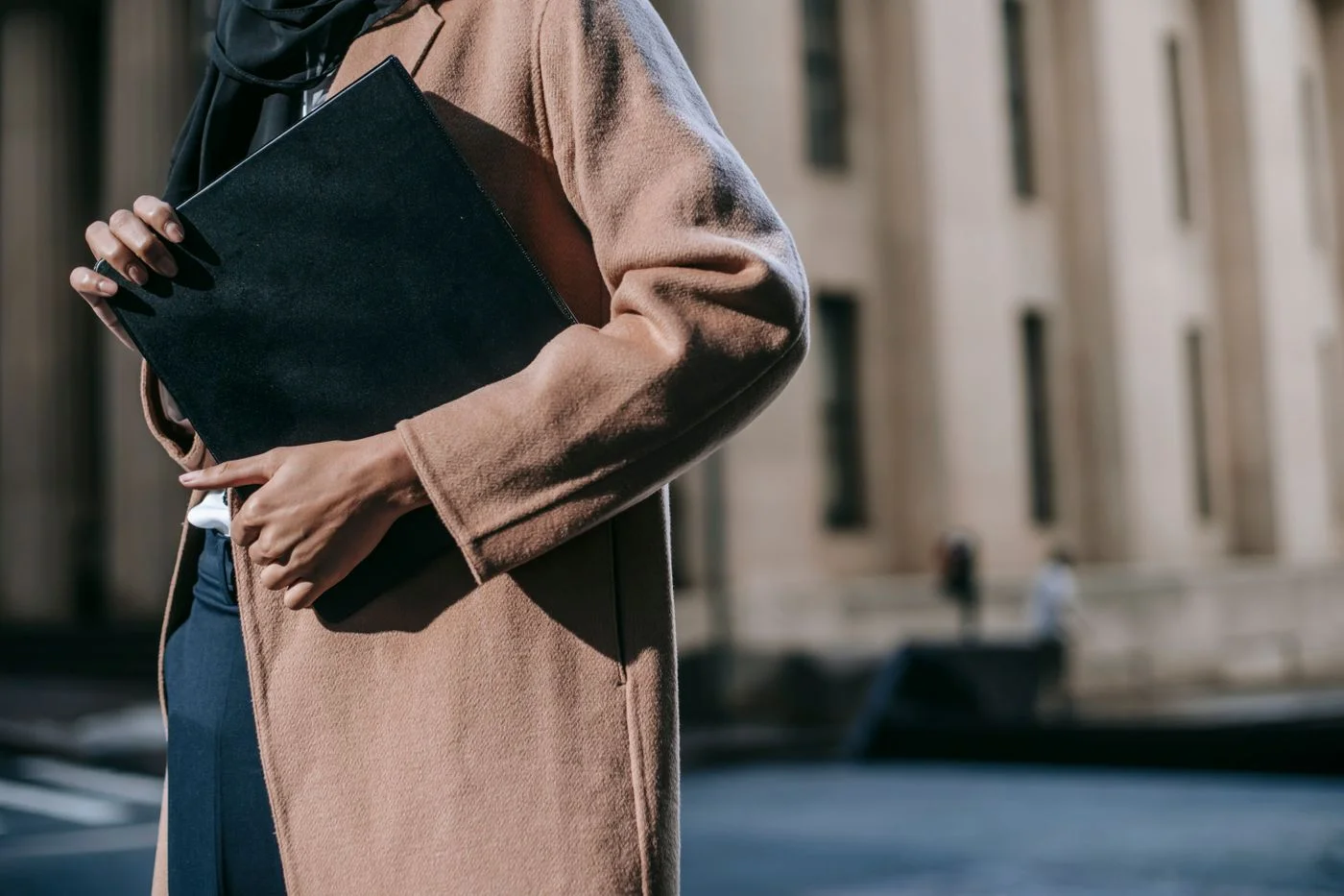 A person in a brown coat carrying a folder of documents outdoors.
