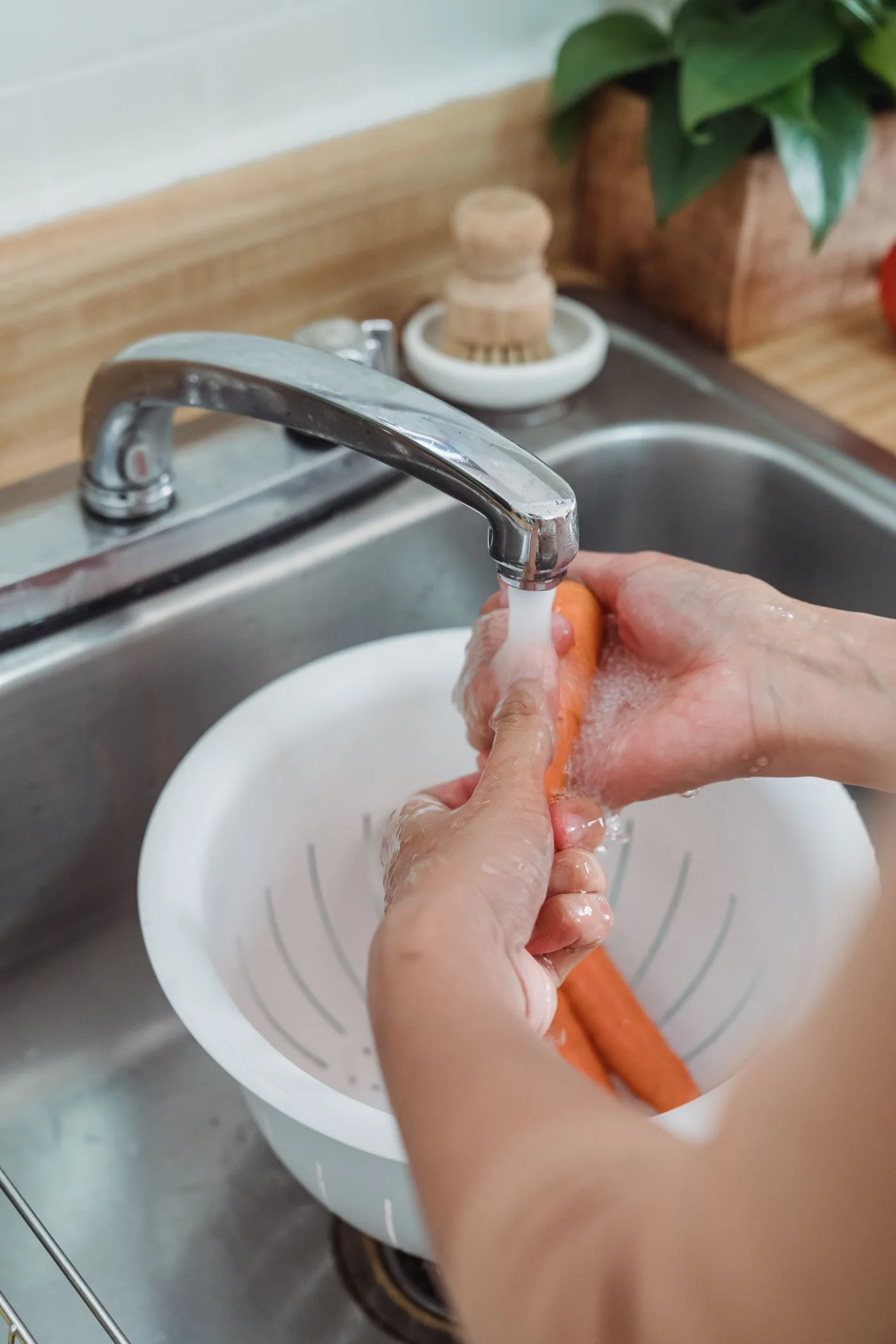 Hands washing carrots in a colander beneath a kitchen faucet.