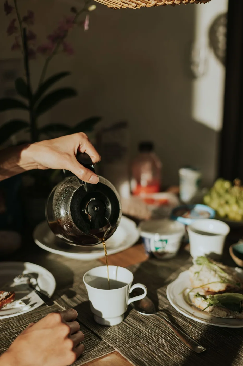 Coffee being poured from a glass pot into a cup beside a breakfast table